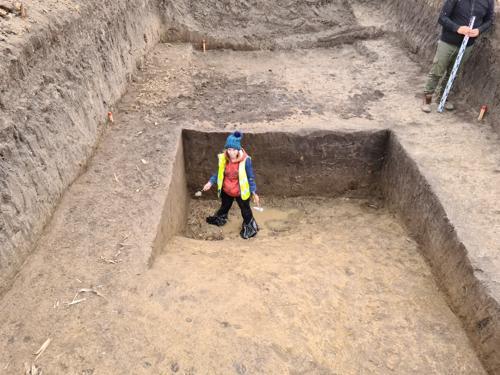 archeologo con cappello blu, felpa rossa e giubbotto ad alta visibilità in piedi in un buco profondo sul sito di scavo