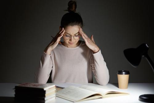donna con gli occhiali seduta a una scrivania che sembra stressata, con libri, tazza di caffè e lampada