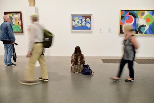 donna seduta per terra in una galleria guarda un dipinto mentre persone sfocate si muovono intorno a lei