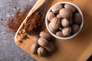 Una foto che mostra una vista dall'alto di una piccola ciotola bianca di semi di noce moscata su una tavola insieme a un cucchiaino di spezia accanto ad altri semi di noce moscata. La tavola è appoggiata su una superficie di pietra e c'è della polvere di noce moscata versata sul bordo.