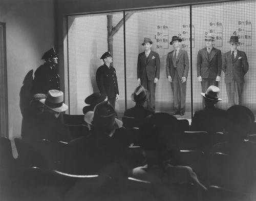 foto in bianco e nero di testimoni seduti di fronte a una fila di 4 uomini con due agenti di polizia presenti