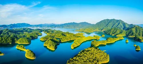Una foto aerea del lago Qiandao, che mostra varie piccole isole densamente alberate nell'immensa distesa di acqua blu. La foto è stata scattata in una giornata luminosa, che conferisce alle isole una leggera tonalità giallastra. Sullo sfondo, una catena montuosa si estende da destra a sinistra, dove svanisce nella distanza.