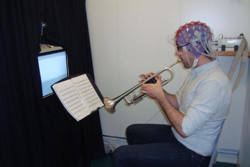 uomo seduto su una sedia che suona la tromba da uno spartito musicale indossando un cappello EEG