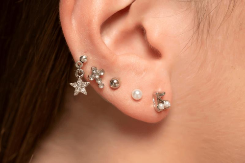 Fotografia di un orecchio con cinque piercing d'argento, da sinistra a destra: una stella pendente, uno spillo a croce, uno spillo d'argento, una perla e una ciliegia con perle