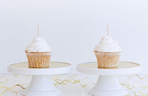 Fotografia di due cupcakes identici con glassa bianca e uno stuzzicadenti che sporge, affiancati su supporti bianchi identici