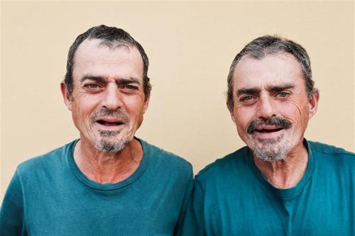 Due uomini gemelli più anziani stanno uno accanto all'altro e sorridono alla fotocamera. Indossano entrambi magliette verdi identiche e hanno lo stesso taglio di capelli corto e barbe curate.