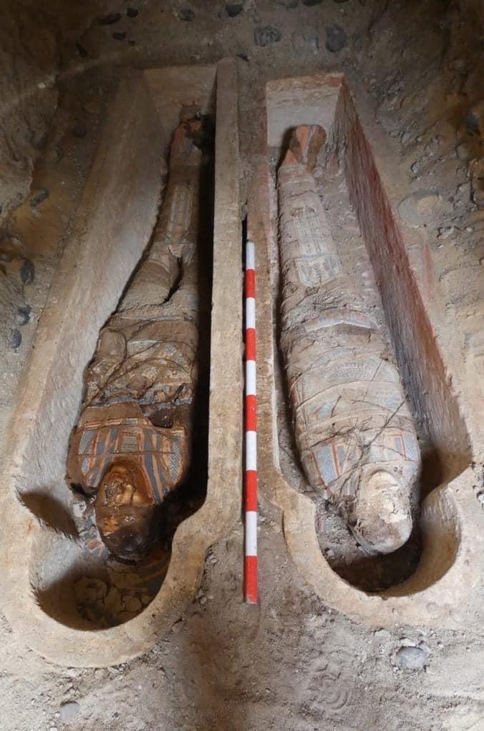 Una foto di due delle mummie recentemente scoperte a Oxyrhynchus. Le mummie sono disposte in due sarcofagi poco profondi a forma di fossa che si trovano uno accanto all'altro. I lati delle fosse sono circondati da cumuli di detriti scavati.