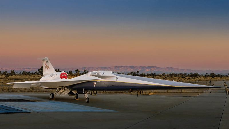 NASA’s X-59 quiet supersonic research aircraft sits on the apron outside Lockheed Martin’s Skunk Works facility at dawn in Palmdale, California. It has a thin long nose that takes a third of its length and a single engine mounted on top below the tail