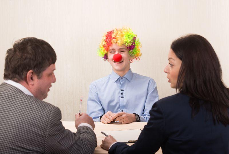 un uomo con una parrucca da clown e un naso rosso sorride mentre i colloquiatori, un uomo e una donna in abito, si girano per discutere di lui