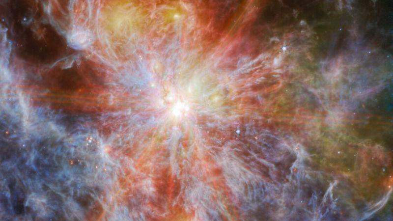 Una giovane stella luminosa all'interno di una nebulosa colorata. La stella è identificabile come il punto più luminoso nell'immagine, circondata da sei grandi raggi di luce che attraversano l'immagine. È possibile vedere anche altri punti luminosi nelle nuvole, che sono mostrate in grande dettaglio come strati di filamenti colorati.