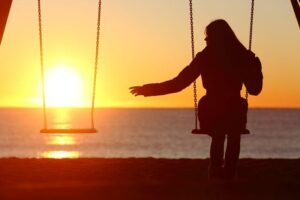Persona in silhoutte seduta su un'altalena al tramonto, tendendo la mano verso un'altalena vuota accanto a loro