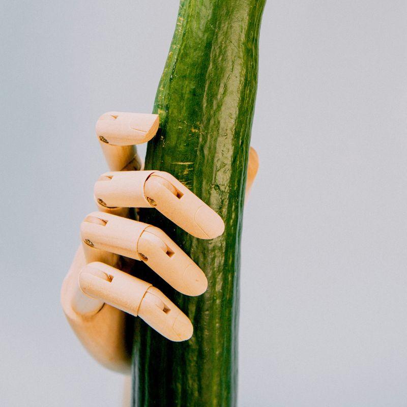 Fotografia di una mano di manichino di legno che tiene un cetriolo su uno sfondo grigio chiaro