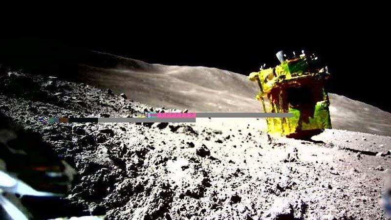 SLIM come si trova attualmente sulla superficie della Luna, visto da uno dei suoi rover. Il lander dorato è capovolto con i razzi puntati verso l'alto e i pannelli solari in ombra.