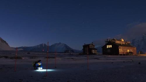 Campionamento della neve a Ny-Ålesund, Svalbard. Notte buia. Artico. Struttura di ricerca sullo sfondo. Persona che campiona la neve con la luce.