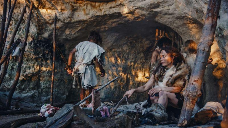 prehistoric-looking-woman-poking-a-fire-with-a-stick-sat-by-a-cave-other-individuals-in-the-background-m-1.jpg
