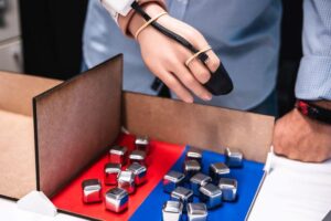 cubi metallici su sfondi blu e rossi su un tavolo; vista della mano protesica di un uomo con un dispositivo nero collegato sul retro della mano e sopra l'indice e il medio