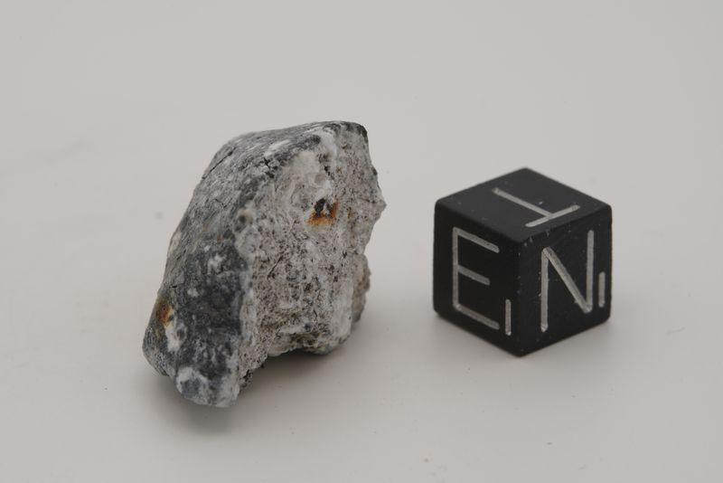 Una rara meteorite aubrite: una roccia grigia con alcune macchie marroni è visibile accanto a un piccolo cubo nero