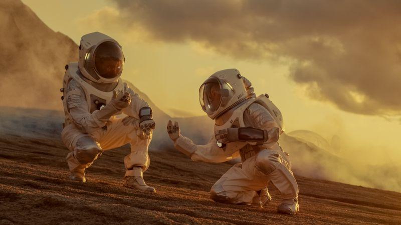 Due astronauti su una collina sabbiosa marrone-arancione, inginocchiati per raccogliere qualcosa