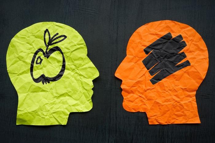 arte concettuale per illustrare l'afantasia; due sagome di carta di una testa umana si fronteggiano su uno sfondo scuro. Quella a sinistra è verde lime e ha un disegno di una mela al suo interno in penna nera; quella a destra è arancione e ha un graffio nero dove dovrebbe esserci la mela.