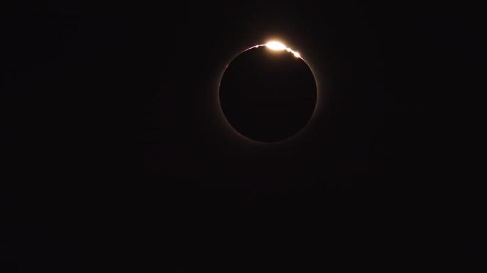 Una foto di un'eclissi solare che mostra l'anello luminoso del sole oscurato dalla luna sospeso nell'oscurità. In cima all'anello luminoso c'è una macchia di luce che rappresenta le Perle di Baily.