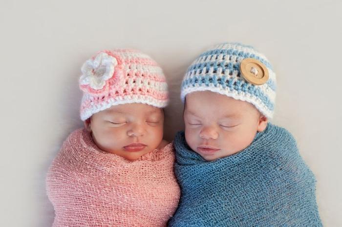due bambini che dormono uno accanto all'altro, uno avvolto in una coperta rosa indossando un cappello all'uncinetto rosa e bianco, l'altro avvolto in una coperta blu indossando un cappello all'uncinetto blu e bianco