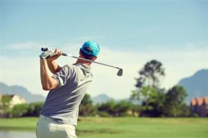 Una foto di un uomo che ha appena tirato un colpo di golf in piedi su un campo da golf. La posizione dell'uomo mostra il bastone sopra la sua spalla sinistra, suggerendo che abbia appena tirato dopo aver colpito la palla. Indossa una berretta blu chiaro, una maglietta grigia e pantaloni bianchi. C'è uno stagno sfocato a sinistra dello scatto e c'è una fila di alberi sullo sfondo.