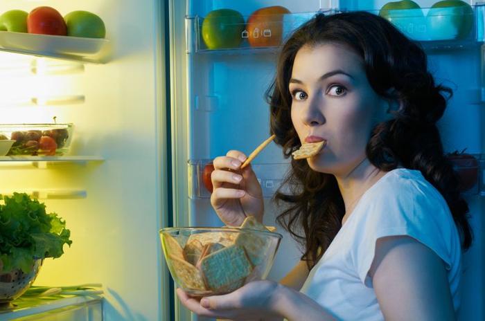 donna mora sembra scioccata alla telecamera dopo essere stata sorpresa a mangiare una ciotola di crackers dal frigorifero; ha un cracker in bocca