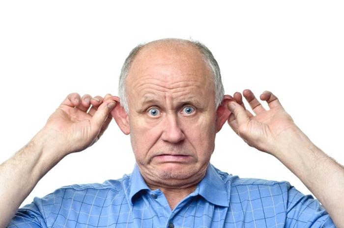 Fotografia di un uomo anziano con una camicia blu che si tira le orecchie e fa una faccia preoccupata