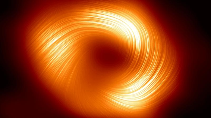 Questa immagine mostra la vista polarizzata del buco nero della Via Lattea. Le linee sovrapposte a questa immagine segnano l'orientamento della polarizzazione, che è legato al campo magnetico attorno all'ombra del buco nero. Una struttura a forma di ciambella luminosa con un buco scuro (il buco nero) è vista coperta da linee che la avvolgono ma non completamente.