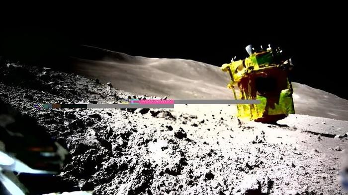SLIM come si trova attualmente sulla superficie della Luna, visto da uno dei suoi rover. Il lander dorato è di lato con i razzi puntati verso l'alto e i pannelli solari in ombra