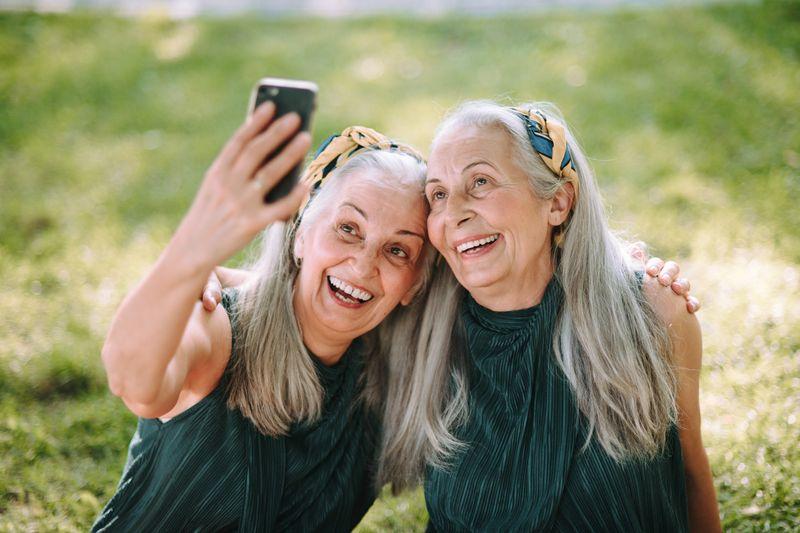 due donne anziane, gemelle identiche, che si fanno un selfie insieme; hanno lunghi capelli grigi e indossano top senza maniche verdi e fasce per capelli verdi e arancioni; sembrano essere sedute in un campo.