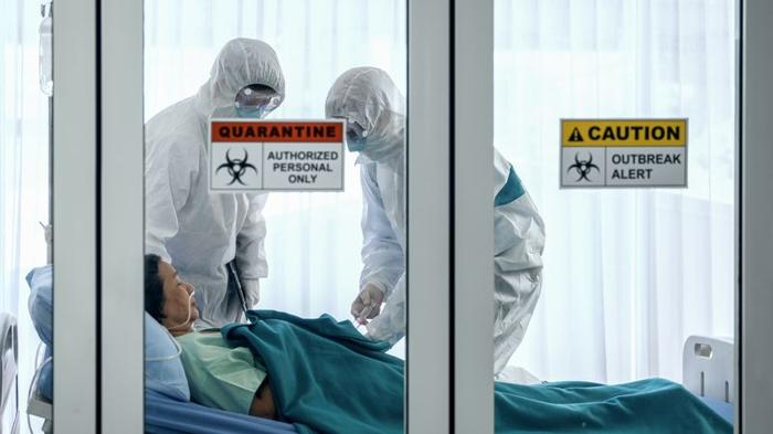 Due persone in tute protettive bianche e mascherine in piedi su una persona in un letto d'ospedale con tubi nel naso, la porta di vetro ha segnali di quarantena