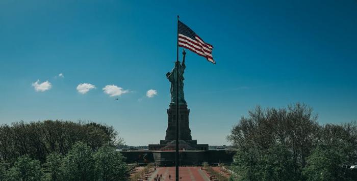 Una bandiera americana sventola davanti alla Statua della Libertà a New York City con un cielo blu brillante.