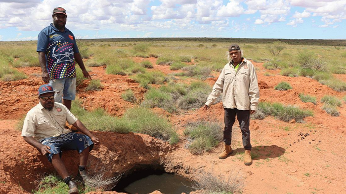 Tre uomini stanno nel deserto vicino a un piccolo buco pieno d'acqua. Questi sono i KJ Martu Rangers.