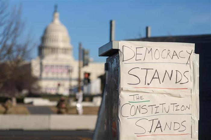 Una foto ravvicinata di un messaggio scritto a mano su carta attaccato a una caratteristica pubblica con Capitol Hill sullo sfondo. Il cartello dice La democrazia resiste. La Costituzione resiste.