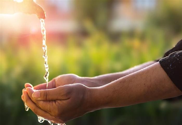 Le mani di un bambino indiano raccolgono acqua mentre scorre da un rubinetto esterno. L'immagine è fortemente saturata dalla luce solare che sfoca l'angolo in alto a sinistra dove si trova il rubinetto. Lo sfondo sfocato mostra un giardino verde lussureggiante o uno spazio aperto simile. 