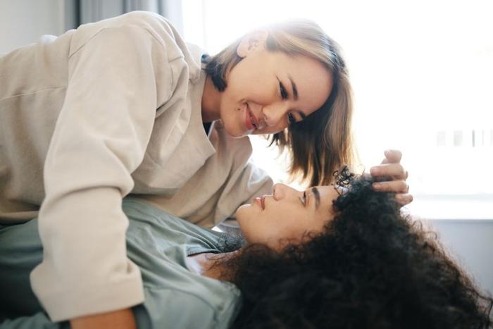 coppia lesbica, una con lunghi capelli ricci scuri e una con capelli più corti con sfumature, distese una sopra l'altra sul letto e che si guardano negli occhi con una finestra luminosa alle spalle