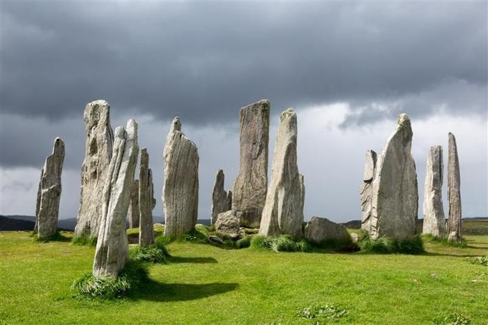Una foto dei menhir sull'Isola di Lewis e Harris, nelle Ebridi Esterne. Le pietre sono disposte in un campo verde lussureggiante e la foto mostra un cielo scuro e tempestoso. Alcune pietre si sono consumate in punte mentre altre conservano ancora una forma più regolare. 