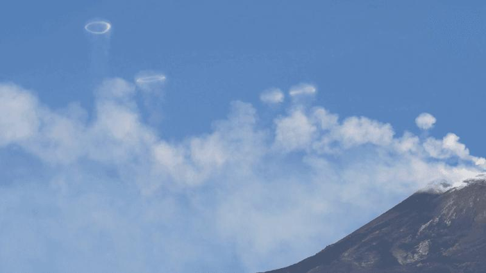 Sul lato destro si trova la cima del Monte Etna e il resto dell'immagine è cielo blu e vapore bianco. Sopra il vapore bianco ci sono anelli perfetti simili a fumo.