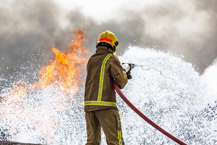 Un pompiere sta con le schiena alla telecamera. Sta spruzzando una schiuma su un incendio che scorre da un tubo rosso.