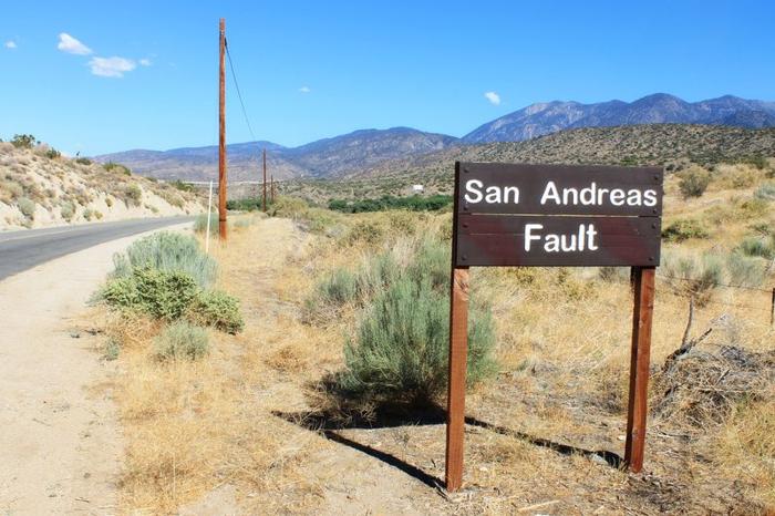 Un cartello che dice 'Faglia di San Andreas', in piedi in un'area secca e erbosa con colline sullo sfondo.