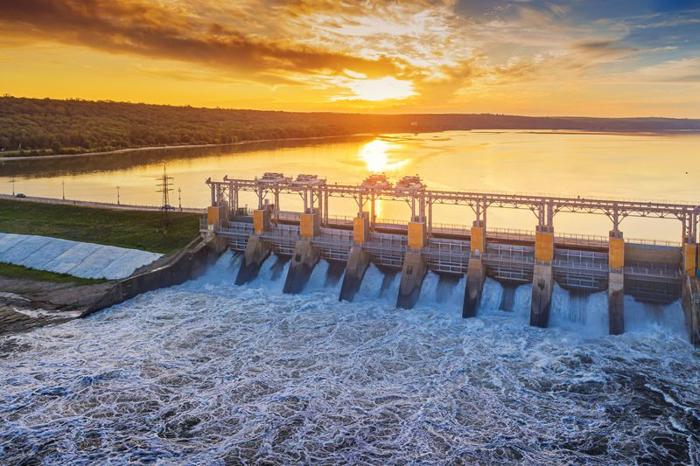 Una foto di una centrale idroelettrica al tramonto. L'acqua scorre attraverso una serie di canali. L'acqua dietro la diga è dorata poiché riflette il sole.