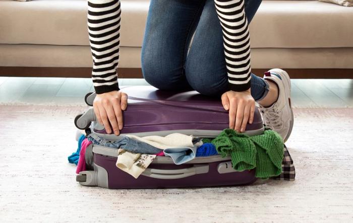 person-wearing-jeans-and-striped-top-kneeling-on-purple-overfilled-suitcase-that-will-not-shut-m-1.jpg