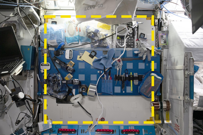 Una foto di una sezione della ISS dove attrezzi e vari altri oggetti sono stati fissati a una parete. C'è un quadrato giallo proiettato nello spazio che è composto da trattini. Una foto di una sezione della ISS dove attrezzi e vari altri oggetti sono stati fissati a una parete. C'è un quadrato giallo proiettato nello spazio che è composto da trattini.