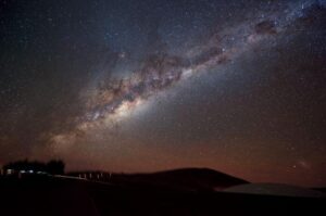 Questa fotografia mostra una vista di taglio della Via Lattea che attraversa il cielo notturno, intrecciata da bande di polvere e gas scuro. Scattata con una fotocamera digitale utilizzando una posa di tre minuti, la fotografia rivela anche un po' di azione a terra. A sinistra, un veicolo con le luci di parcheggio accese si ferma per far scendere un passeggero. Anche se illuminato dalla luce della Via Lattea, il deserto ad alta quota rimane piuttosto scuro. Per illuminare il sentiero verso destra che porta alla rampa di ingresso sotterranea della Residencia, dove il personale e i visitatori alloggiano, il passeggero porta con sé una piccola torcia, vista come una linea luminosa ondulata. In basso a destra, la cupola di vetro sul tetto della Residencia riflette il cielo stellato sopra di essa. Uno dei satelliti galattici della nostra Via Lattea, la Grande Nube di Magellano, è visto sospeso sopra la Residencia nell'angolo in basso a destra.