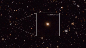 Un cielo nero pieno di galassie alcune abbastanza vicine da mostrare caratteristiche, la maggior parte sono solo punti rossi. Un'immagine ingrandita mostra la galassia in questione. Anche la versione ingrandita non mostra molte caratteristiche oltre a una struttura luminosa sferica