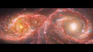 Una versione ingrandita dell'immagine nell'articolo che è descritta come tale. Due galassie a spirale assumono la forma di una maschera colorata con perline che si trova sopra il naso. La galassia a sinistra, IC 2163, è più piccola, occupando poco più di un quarto della vista. La galassia a destra, NGC 2207, occupa metà della vista, con i suoi bracci a spirale che raggiungono i bordi. IC 2163 ha un nucleo arancione brillante, con due bracci a spirale prominenti che ruotano in senso antiorario e diventano più dritti verso le estremità, il lato sinistro si estende quasi fino al bordo. I suoi bracci sono un mix di rosa, bianco e blu, con un'area che assume la forma di una palpebra che appare più bianca. NGC 2207 ha un nucleo molto luminoso. Nel complesso, sembra avere bracci a spirale più grandi e spessi che ruotano in senso antiorario. Questa galassia contiene anche aree blu di formazione stellare più grandi e numerose che spuntano come buchi dai bracci a spirale rosa. Nel mezzo, i bracci delle galassie sembrano sovrapporsi. I bordi mostrano lo sfondo nero dello spazio, inclusa galassie estremamente distanti che sembrano macchie arancioni e rosse, e alcune stelle in primo piano.