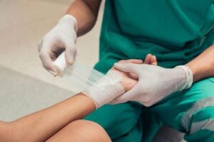 operatore sanitario in uniforme verde e guanti bianchi applica una benda sulla mano del paziente (primo piano)