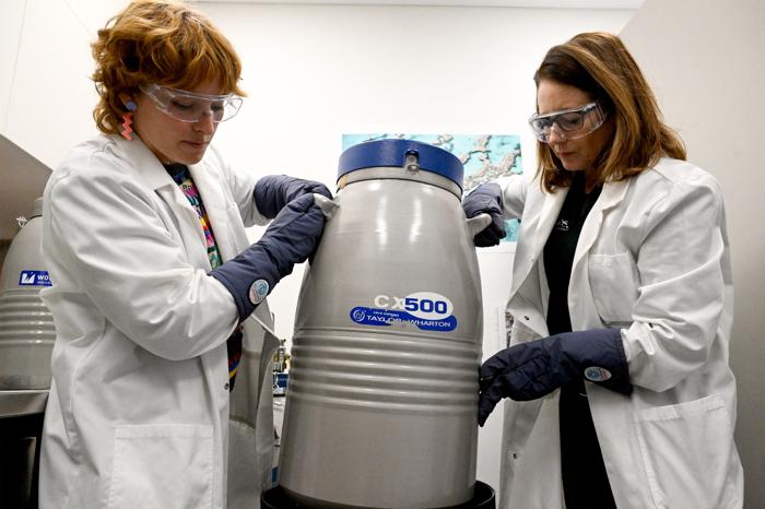 Due donne in camici da laboratorio che spostano un grande serbatoio cilindrico