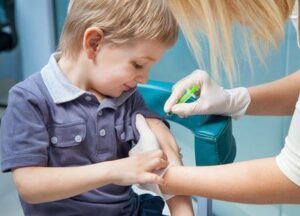 Una foto mostra un bambino piccolo che indossa una camicia di jeans blu con un colletto blu più chiaro mentre viene vaccinato. Il bambino sta guardando il suo braccio sinistro mentre l'infermiera si inclina in avanti per iniettarlo con una piccola siringa verde. Il bambino non sembra affatto preoccupato.